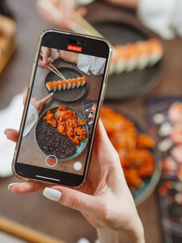 A woman photographing sushi and chicken dish with a smartphone at a restaurant.