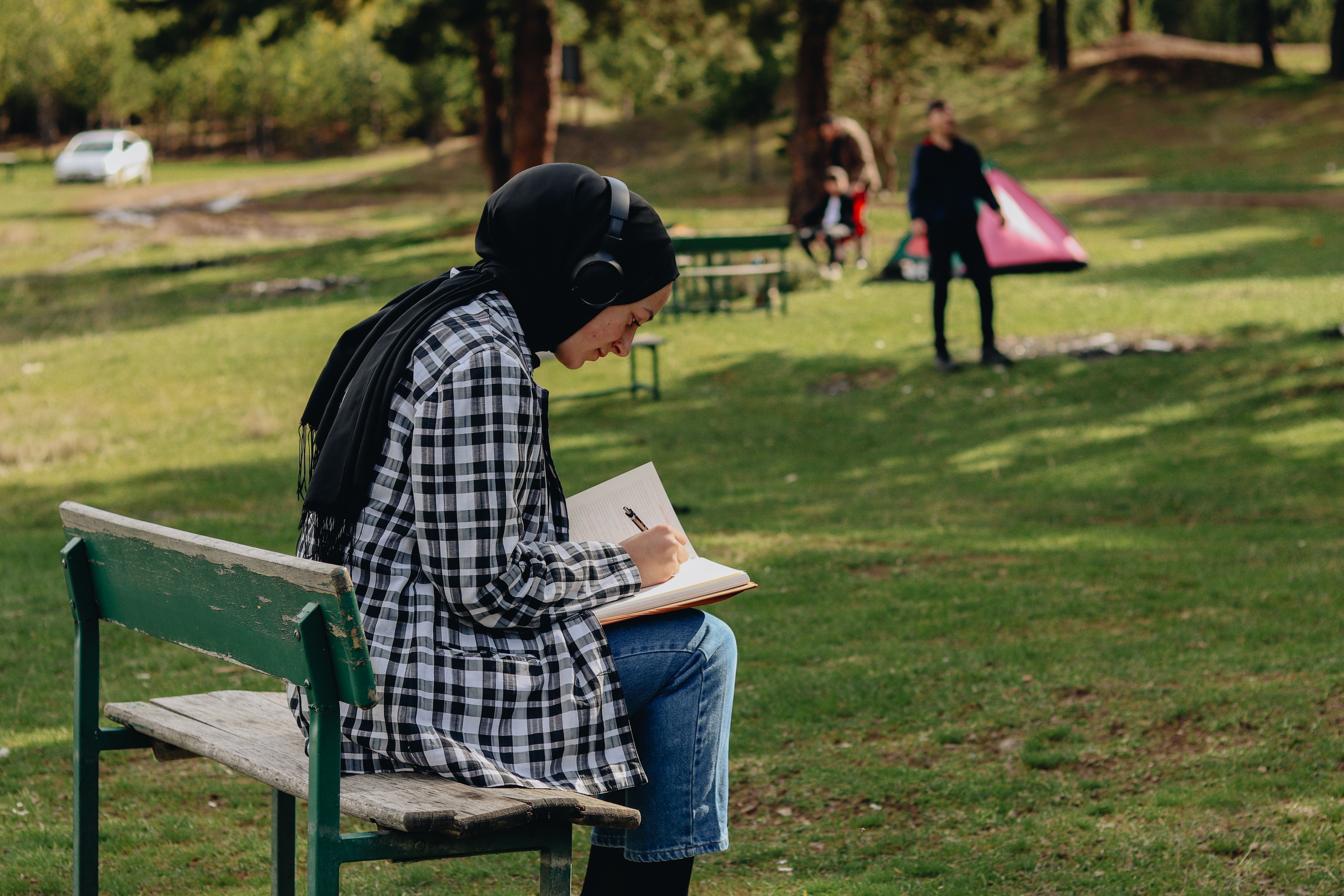 a person sitting on a park bench, writing in a notebook with trees in the background, soft sunlight