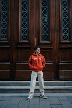 Stylish man in red hoodie posing confidently by a large wooden door in an urban setting.