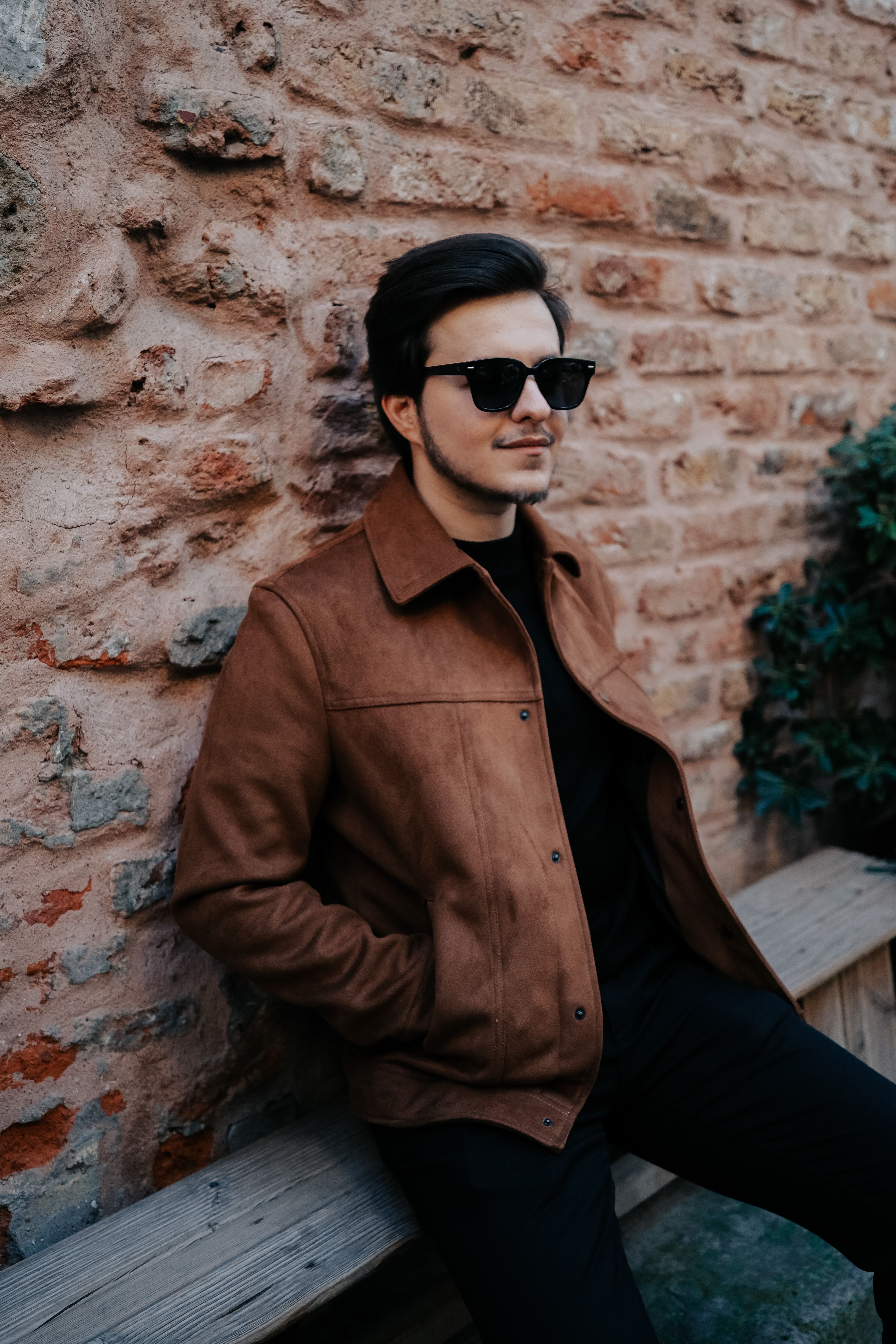 Fashionable man in sunglasses and jacket sits on a bench against a brick wall.