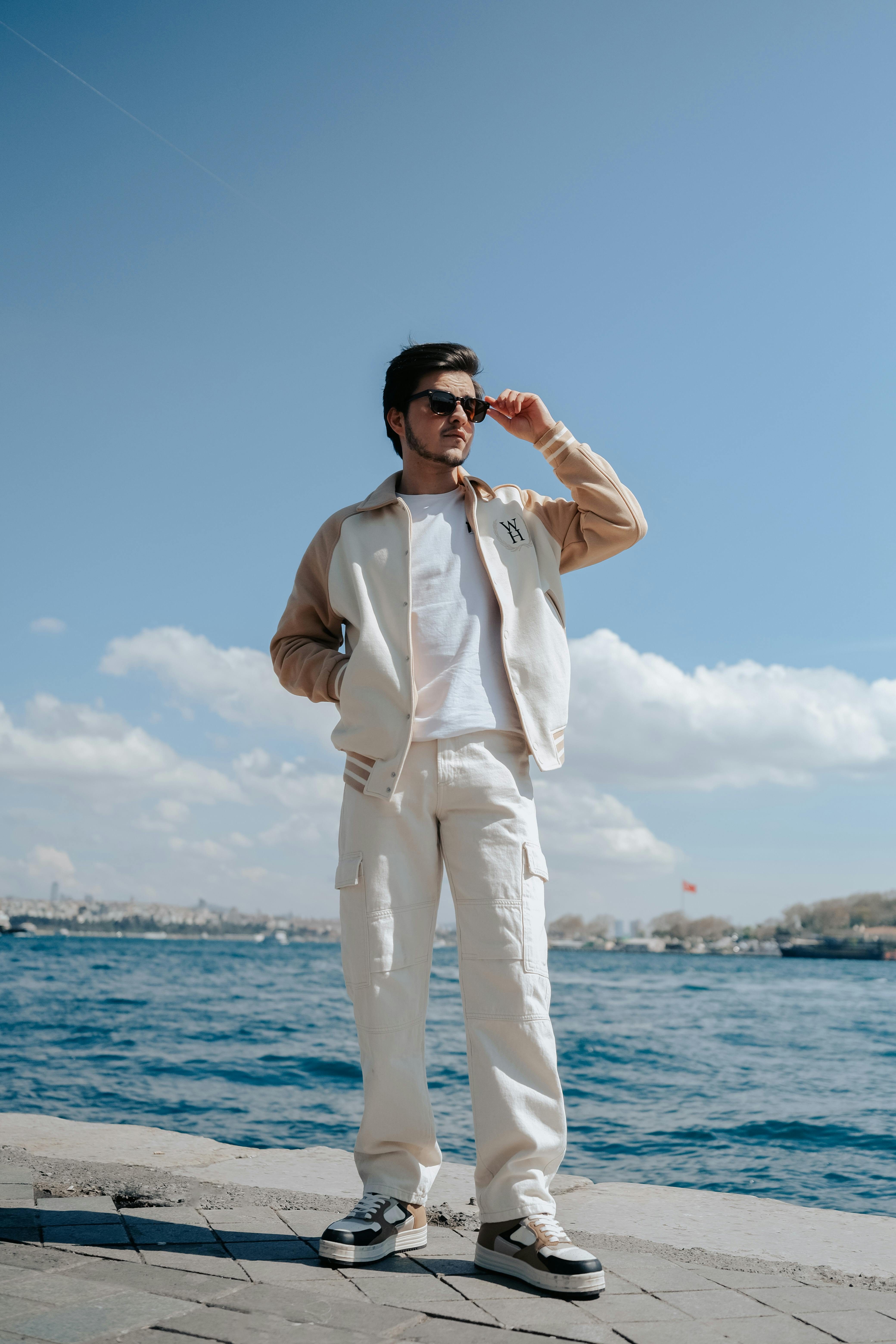Stylish man posing confidently by the seaside, wearing sunglasses and casual attire.