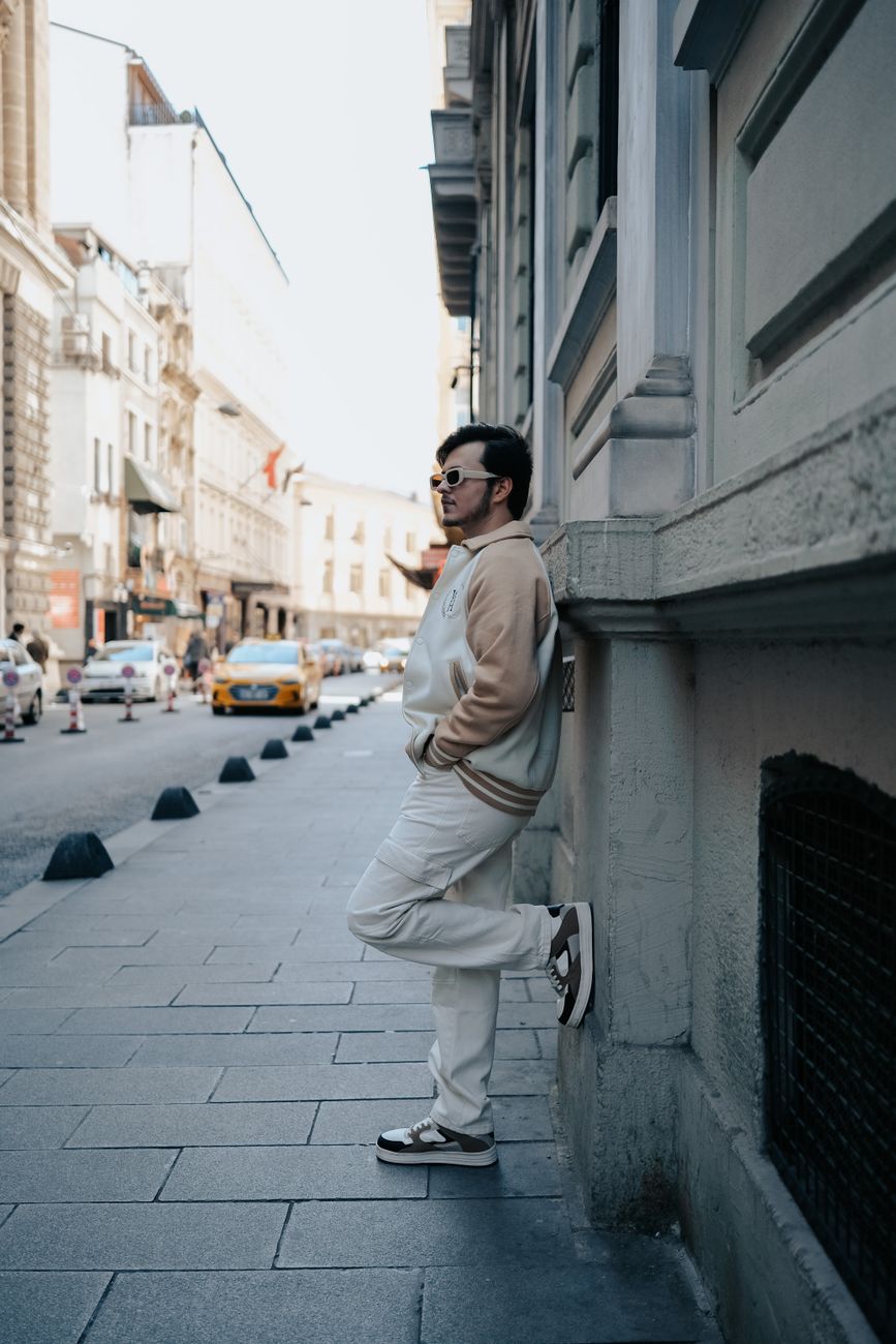 Fashionable man wearing sunglasses leans on a city street wall in a vibrant urban setting.