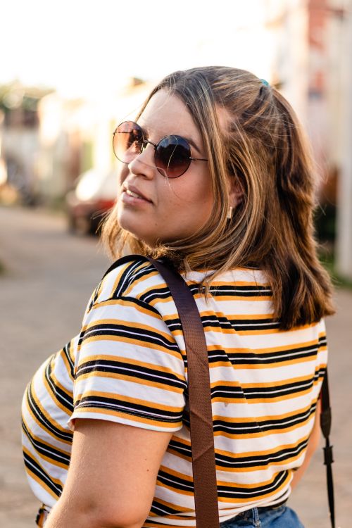 Free A woman with sunglasses poses stylishly outdoors in sunlight, wearing a striped shirt. Stock Photo