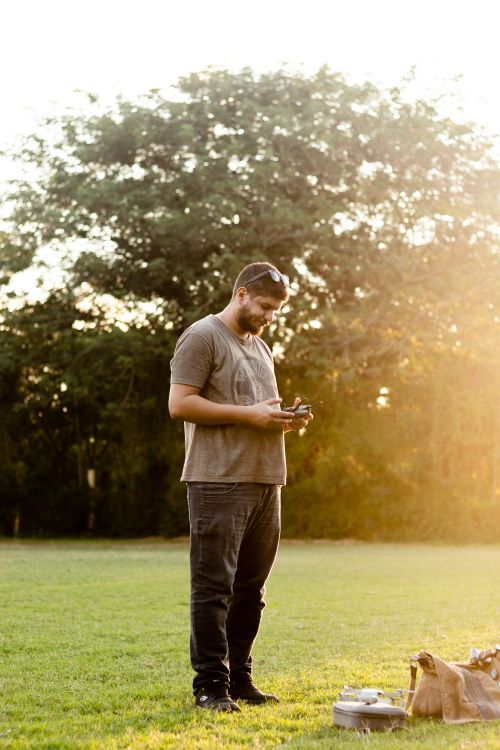 Free Man in brown t-shirt using a remote control to pilot a drone outside in a vibrant park setting. Stock Photo