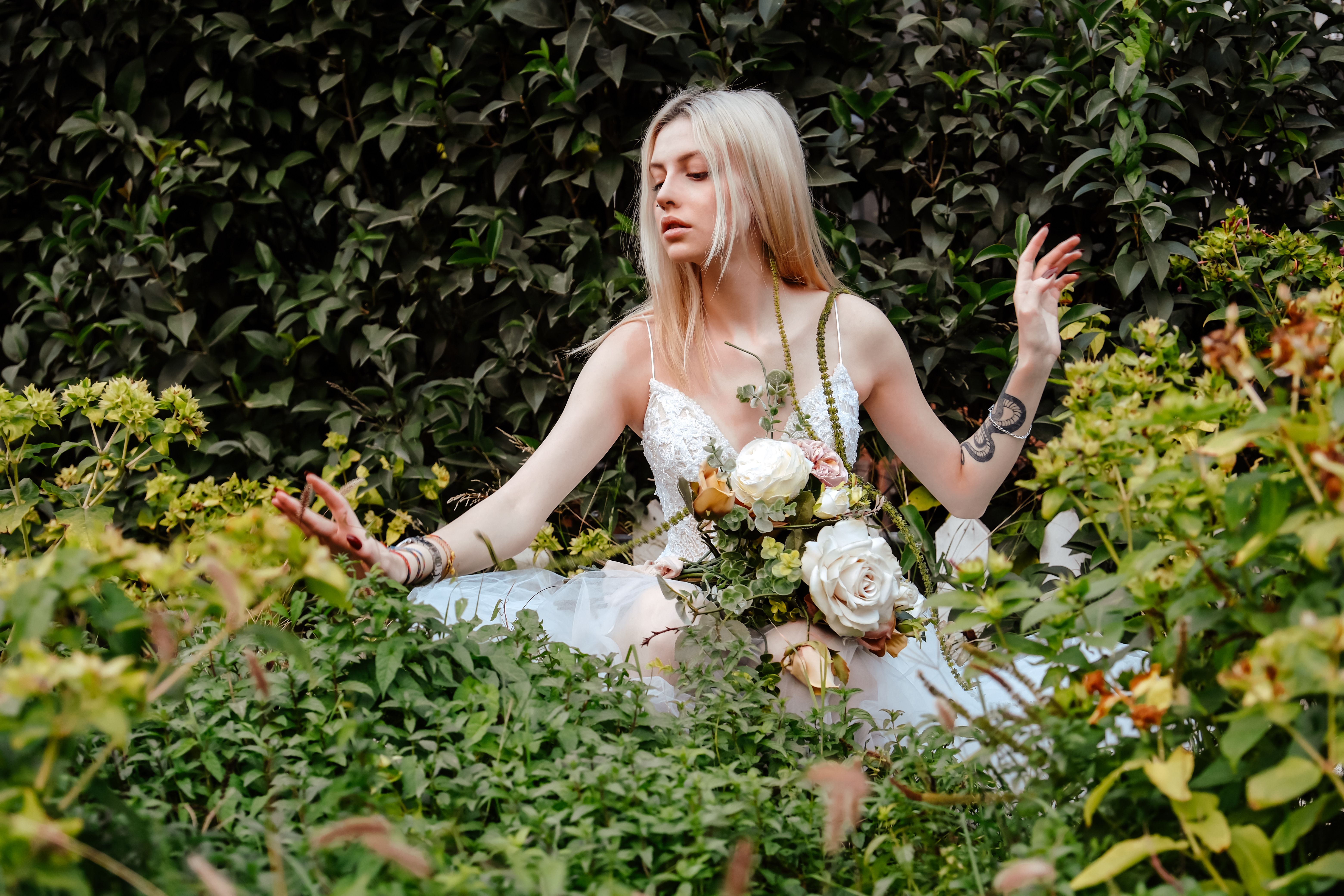 Elegant woman in white dress surrounded by lush garden foliage, holding a bouquet of white roses.