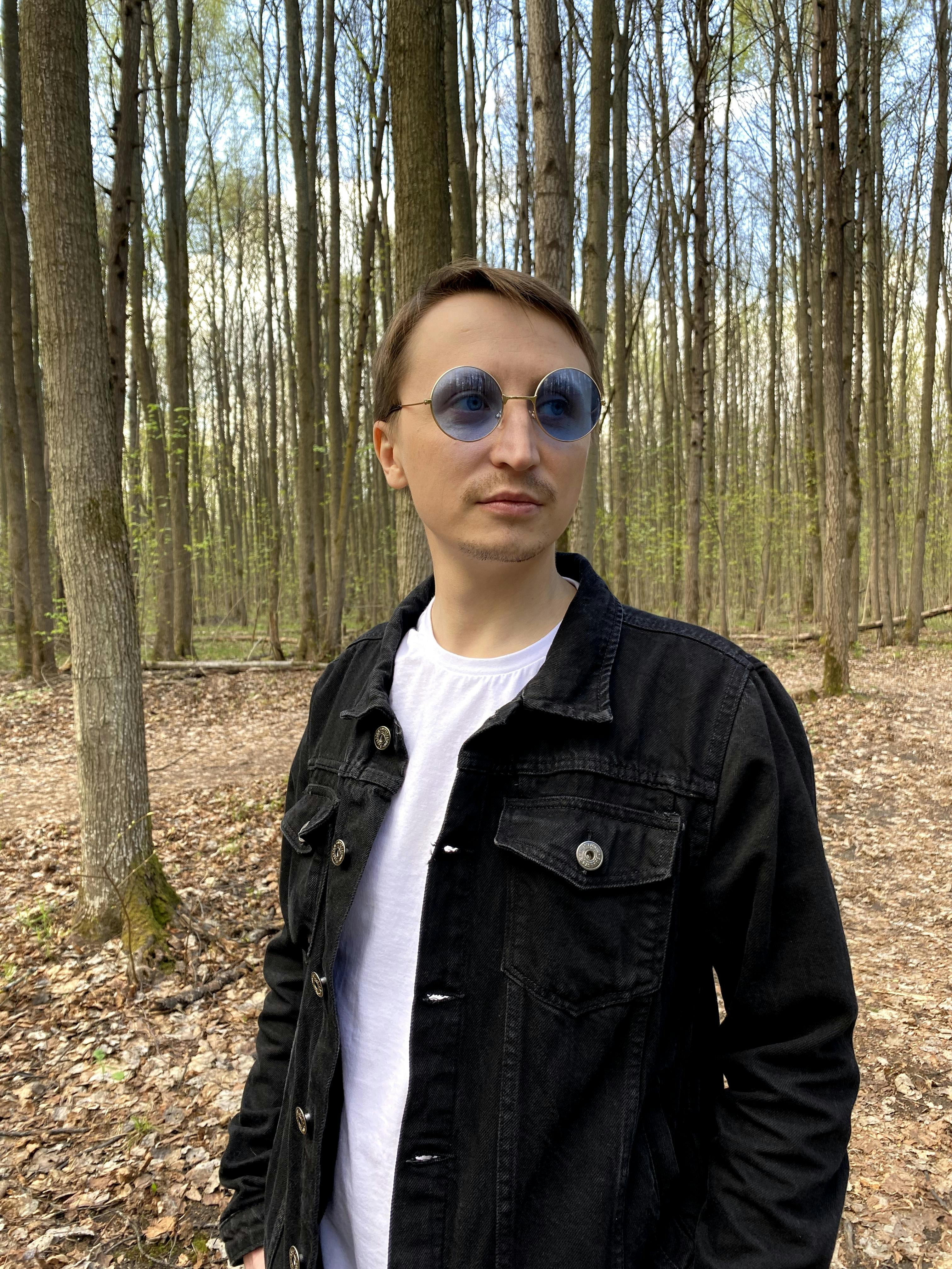 Stylish man in black denim jacket and blue glasses posing in a forest setting.