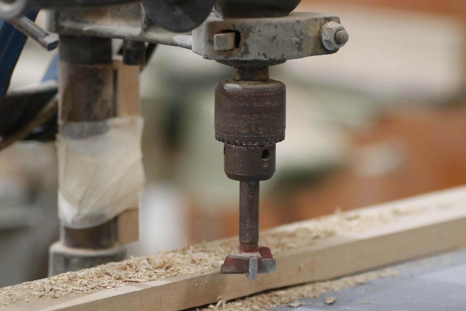 Drill press cutting into a wooden workpiece with wood shavings visible in a workshop