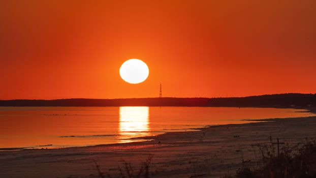 A stunning sunset casts vibrant reflections on calm water, with a sandy shore in the foreground.