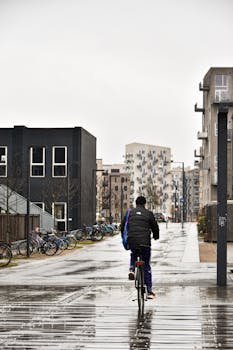 Man cycling in a rainy urban environment with modern buildings.