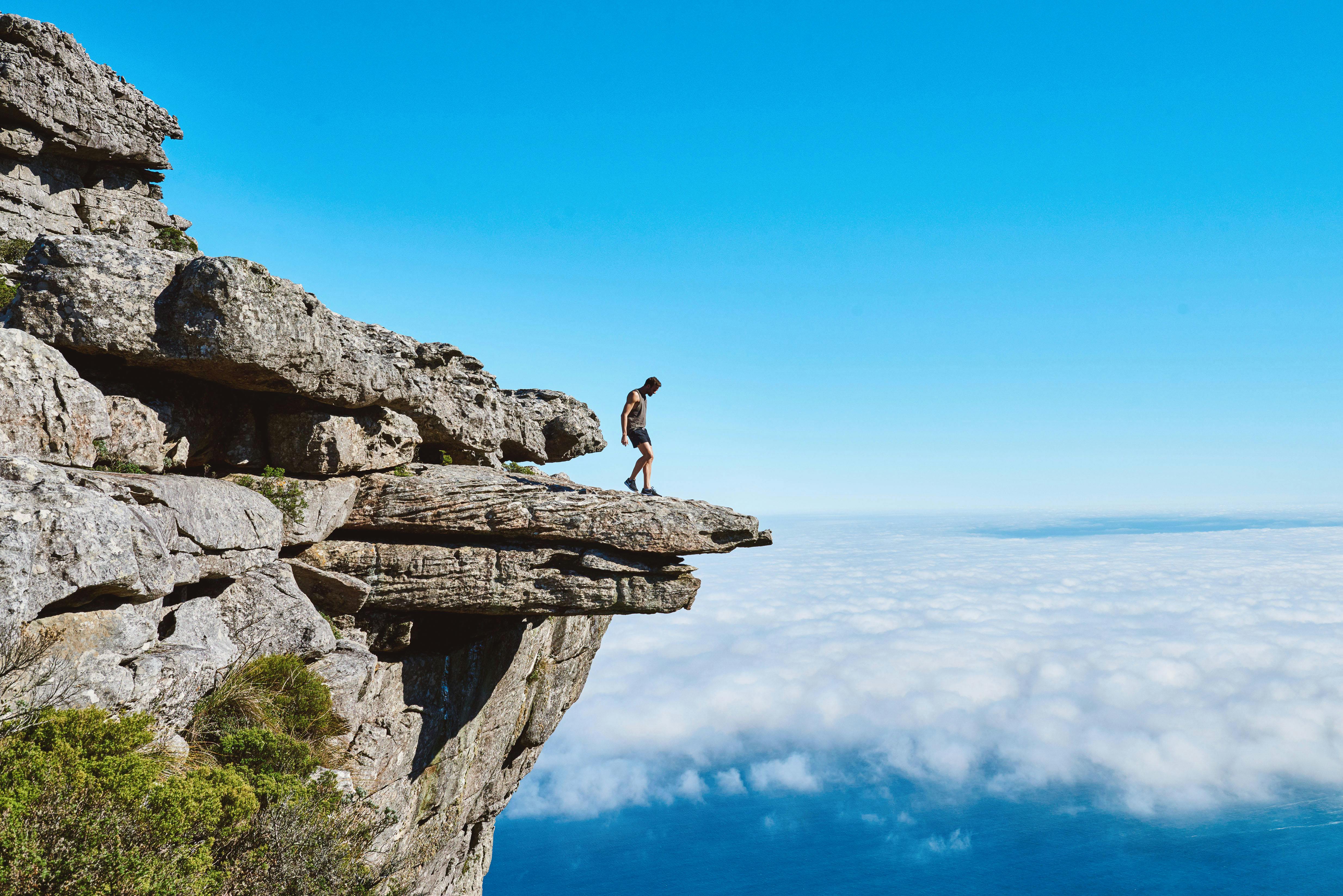 A lone person on a dramatic cliff edge gazing over a sea of clouds under a bright blue sky.