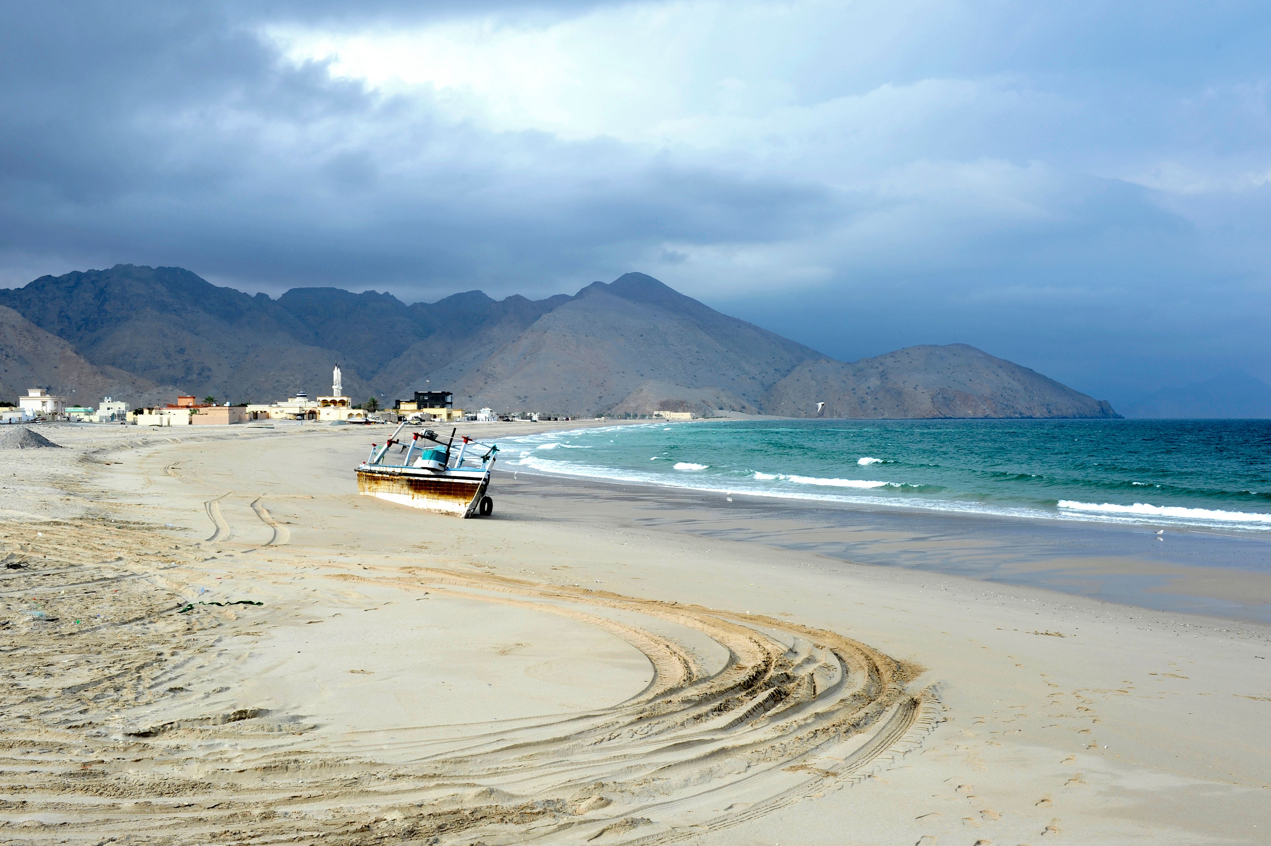 A tranquil beach scene with an abandoned fishing boat, mountains, and ocean waves under a cloudy sky.