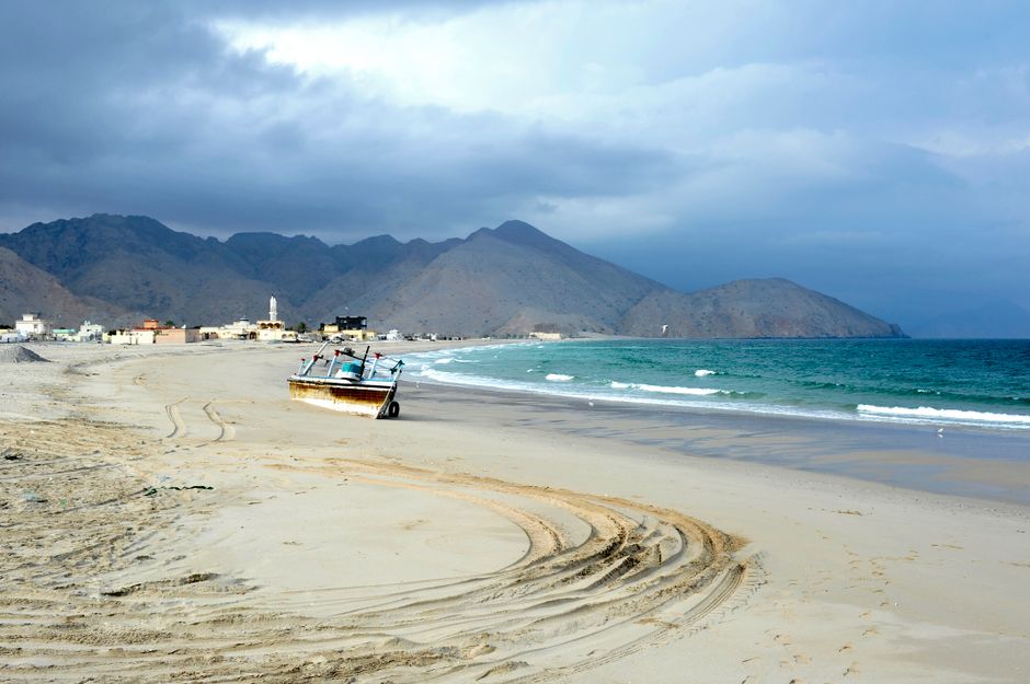 A tranquil beach scene with an abandoned fishing boat, mountains, and ocean waves under a cloudy sky.