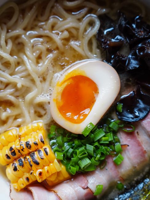 A ceramic bowl of ramen with curly yellow noodles, a halved soft-boiled egg, sliced scallions, nori strips, and slices of pork in a cloudy broth.