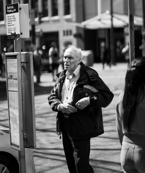 Free Black and white portrait of an elderly man standing at a city bus stop, capturing urban life. Stock Photo