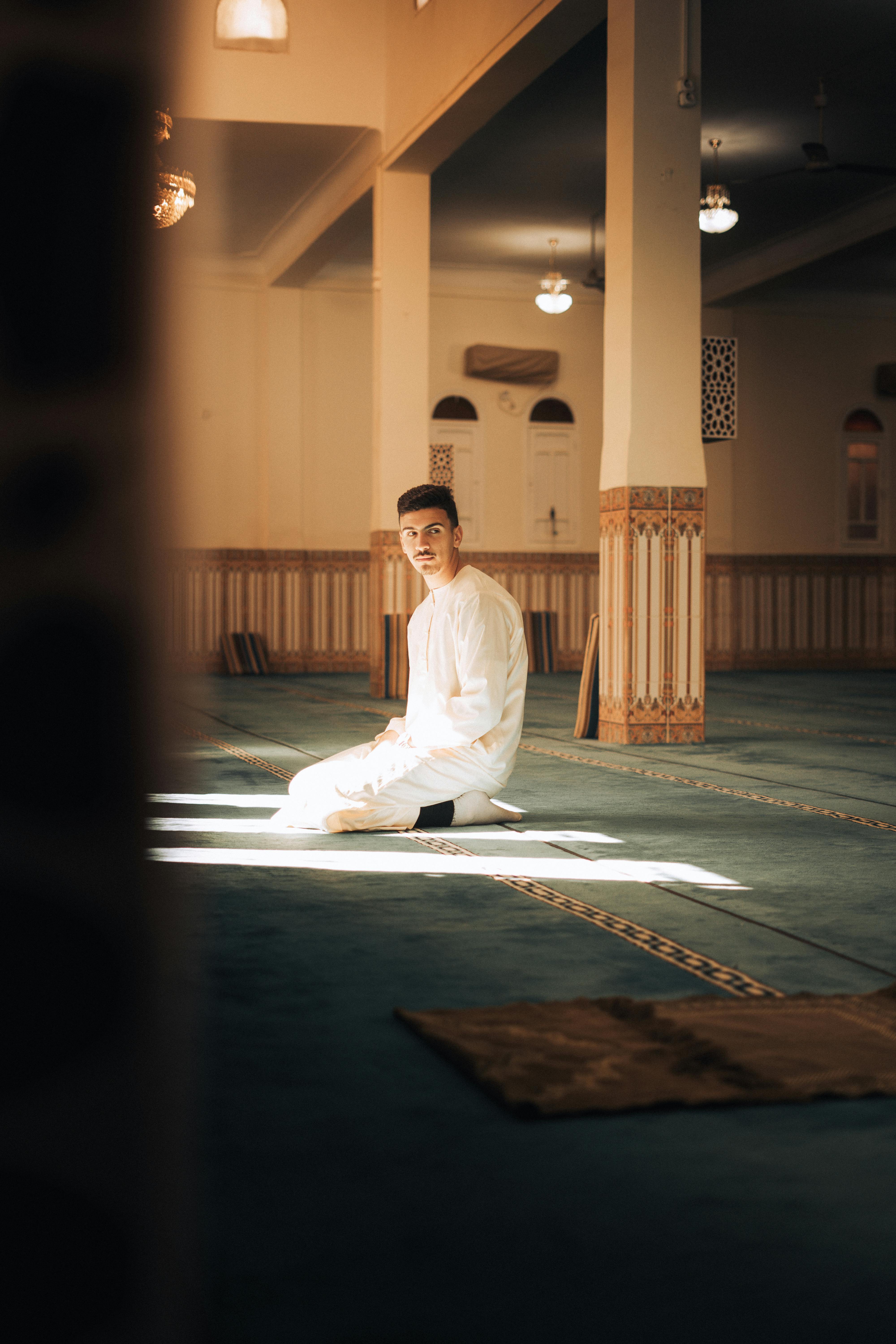 A man in a white gown kneels and prays in a sunlit mosque interior, embracing spirituality and peace.