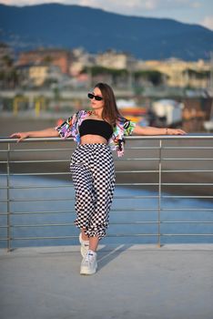 Woman in fashion outfit posing on a bridge with a scenic city background under summer skies.