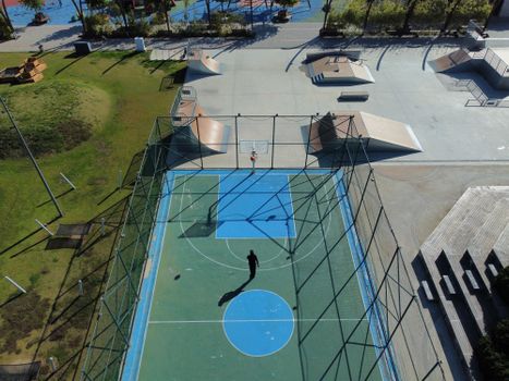 Drone view of a sunlit basketball court and skatepark in an urban area, showcasing sports activity.