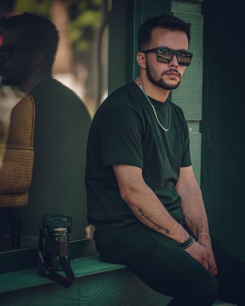 Free Casual man in sunglasses and tattoos sitting by a window with a camera, reflecting urban style. Stock Photo