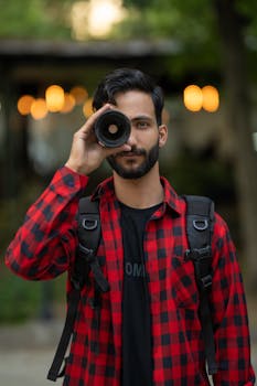 A bearded man wearing a red checkered shirt holds a camera lens like a telescope outdoors.