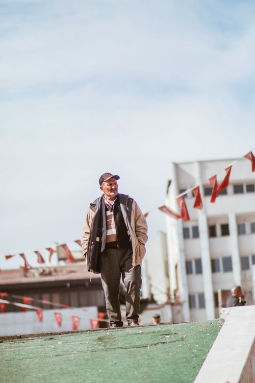 Free Senior man walking outdoors with flags and buildings in the background, capturing leisure and urban life. Stock Photo