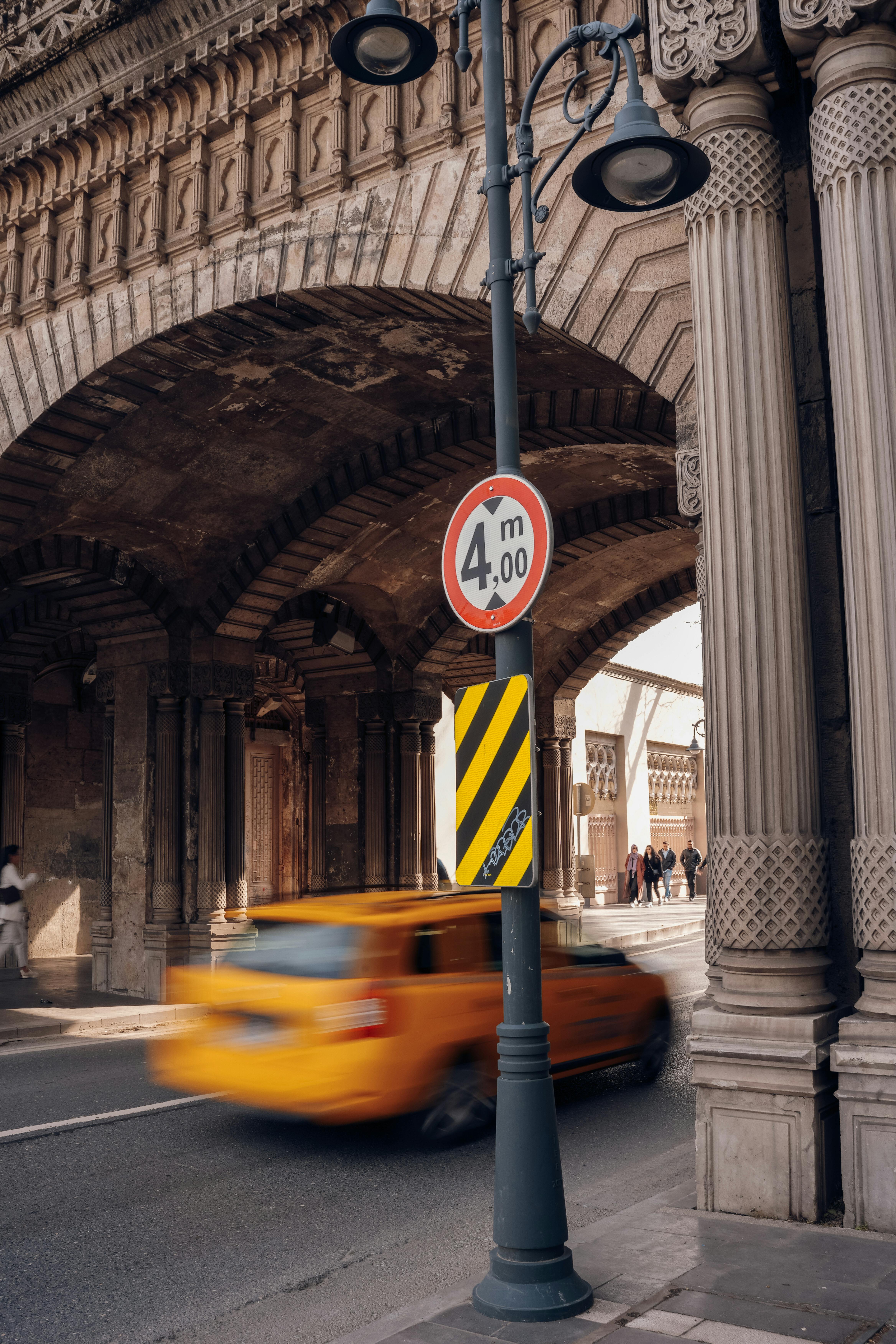 A blurred yellow taxi speeds through a historic urban archway with a prominent height restriction sign.
