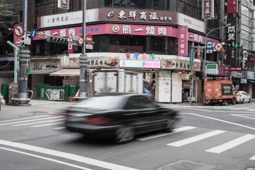 Blurred car moving through a busy Asian city intersection, surrounded by shops and colorful signs.