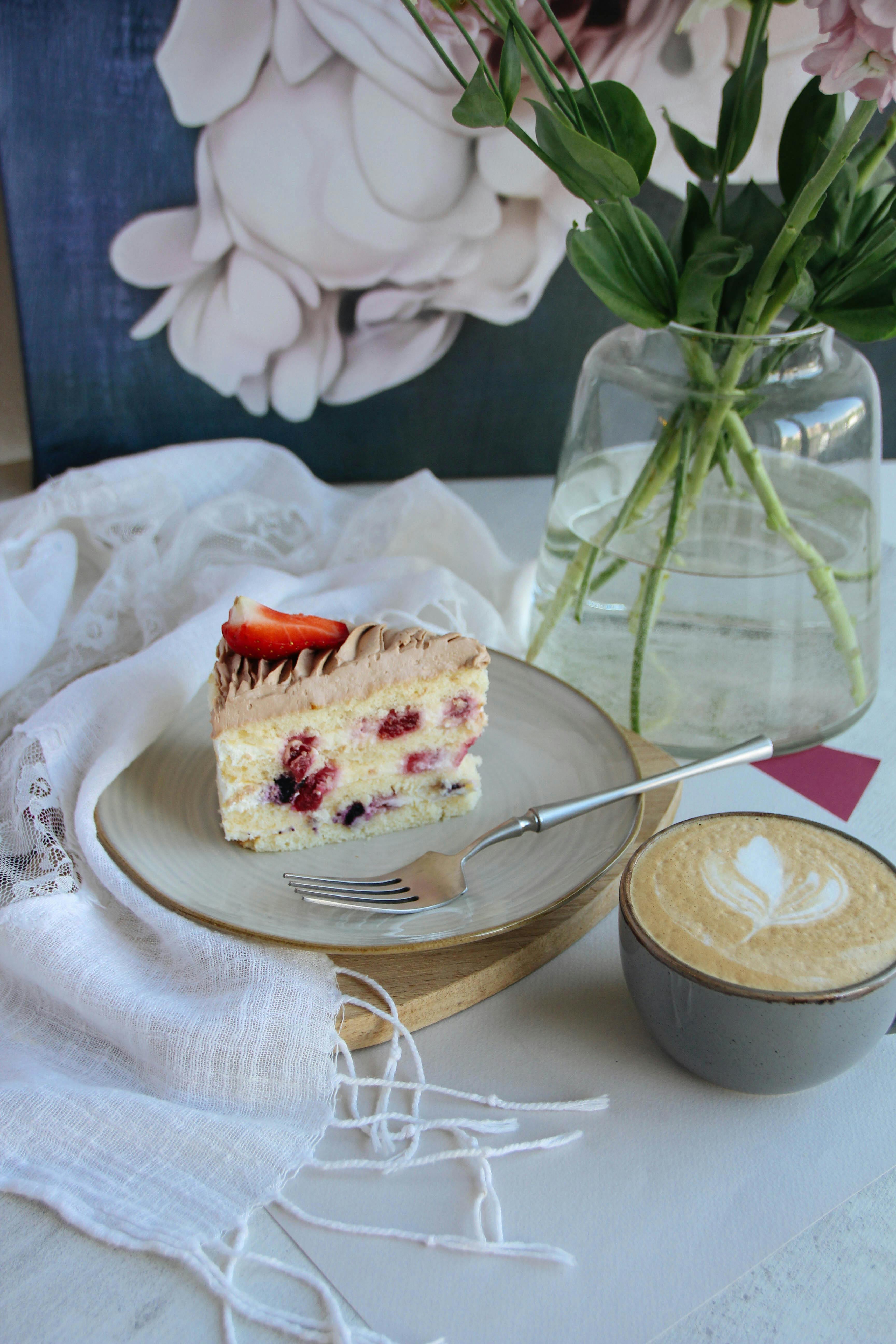 A delicious berry cake slice with coffee beside a vase of flowers on a table.