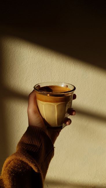 A cozy hand holding a coffee glass in a sunlit room, casting soft shadows.