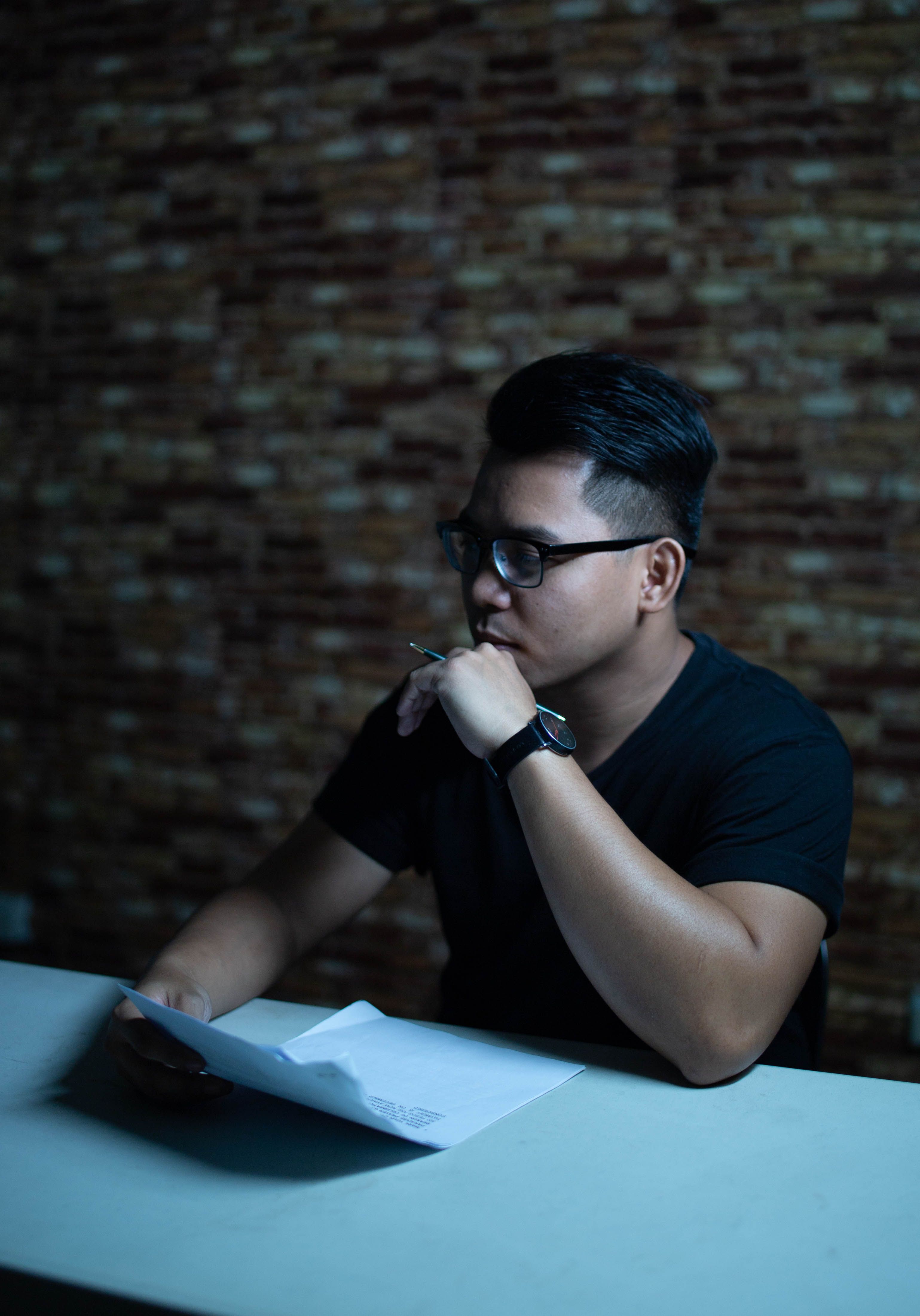 Alt Text: A man with glasses sitting indoors at a wooden table holding a pen while thoughtfully reviewing contract paperwork.