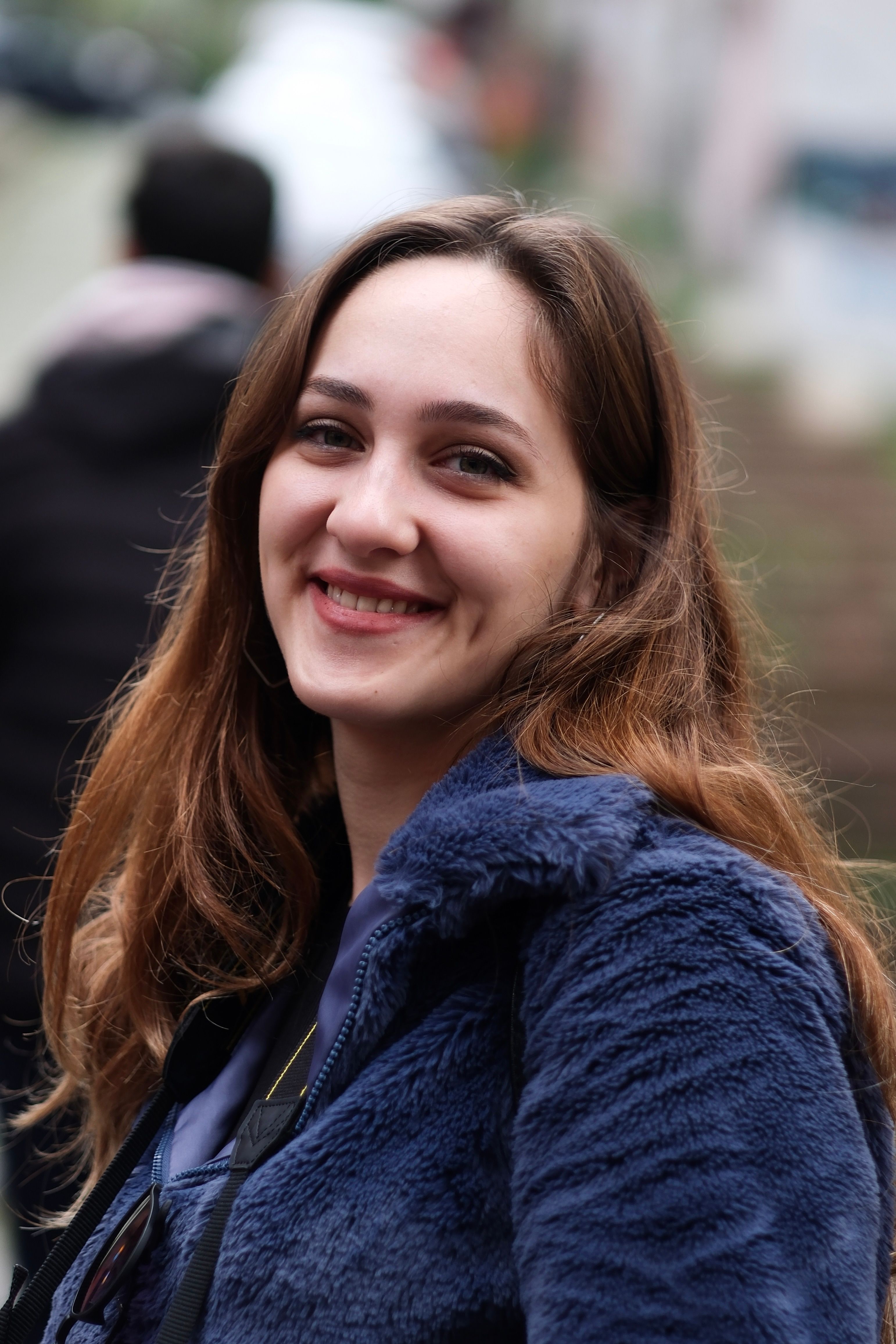 A smiling woman with brown hair poses outdoors in a blue jacket, exuding joy.