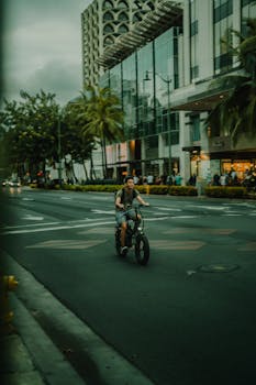 Man biking through bustling Honolulu street with urban cityscape and palm trees in the background.