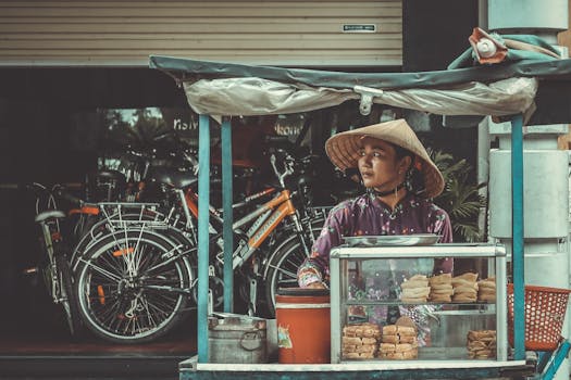 Asian woman selling traditional street food from a cart in a bustling urban area with bicycles in the background.