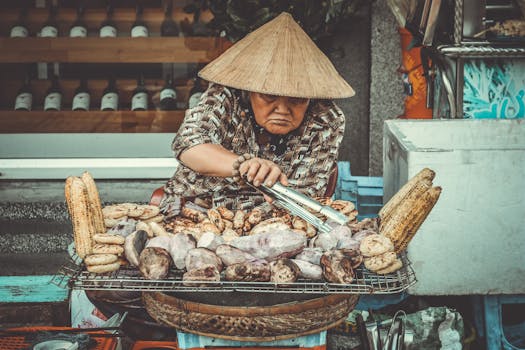 Asian woman grilling corn and sweet potatoes on a street food stand with a traditional hat.