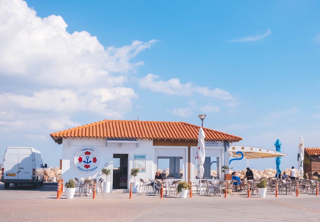 Small white beachside café with terracotta roof and outdoor seating area under blue sky.
