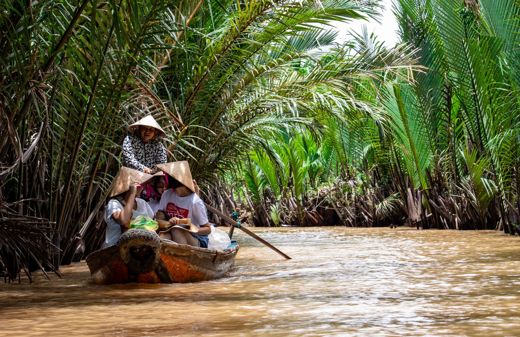 Vietnamese boatwoman rowing three travelers through a lush Mekong Delta palm channel