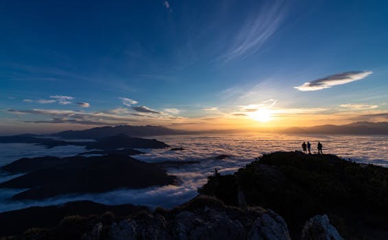 Silhouetted hikers stand on mountain peak at sunrise with clouds below, creating a scenic landscape view.