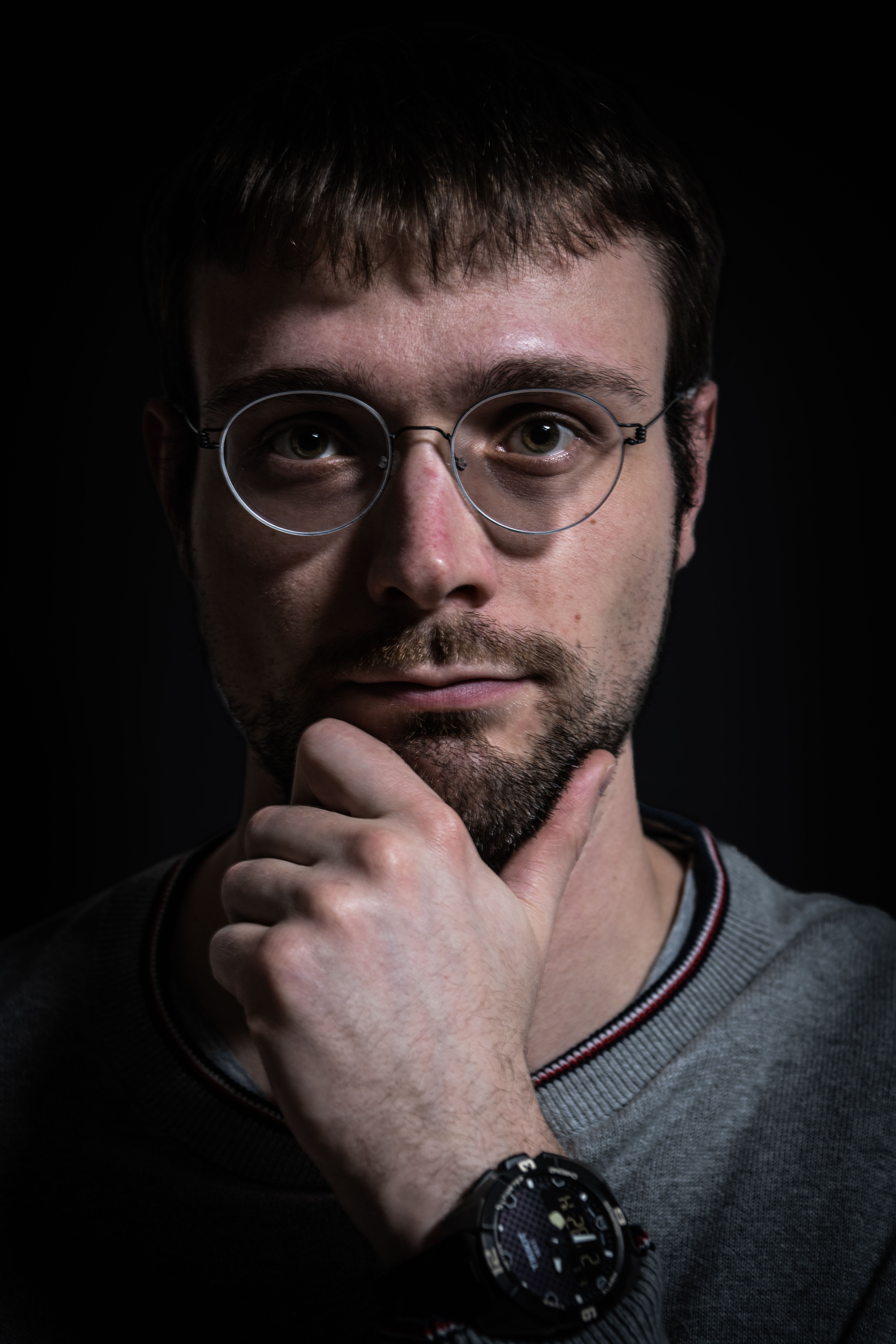 Close-up portrait of a man with eyeglasses and hand on chin, set against a dark background.