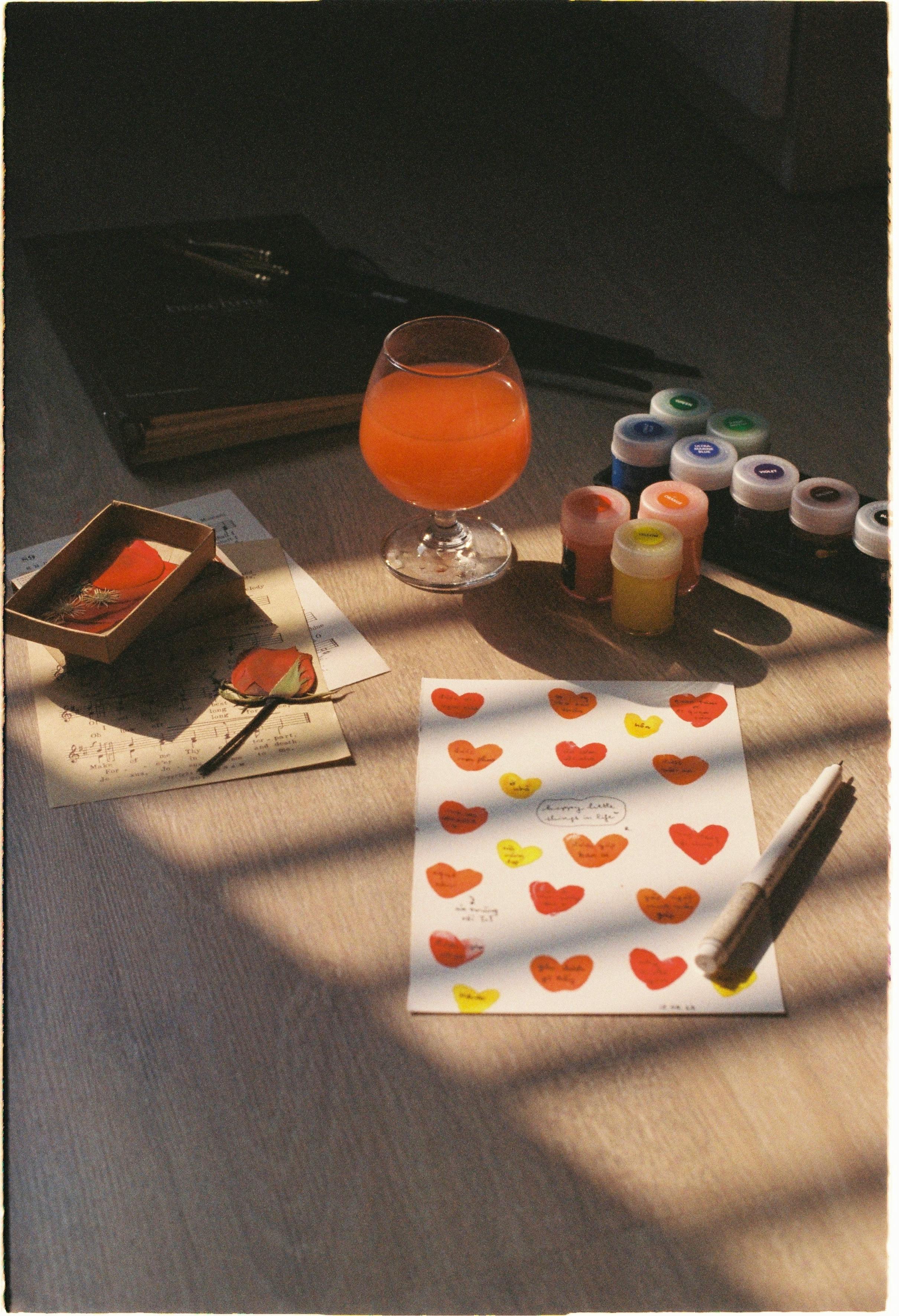 Sunlit art supplies and orange drink on a wooden floor create a warm, creative atmosphere.