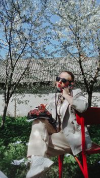 A stylish woman in a vintage suit sits on a red chair using a rotary phone against a backdrop of blossoms.