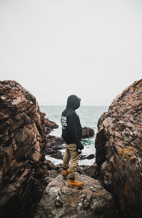 Man in Hoodie Standing between Rocks on Shore