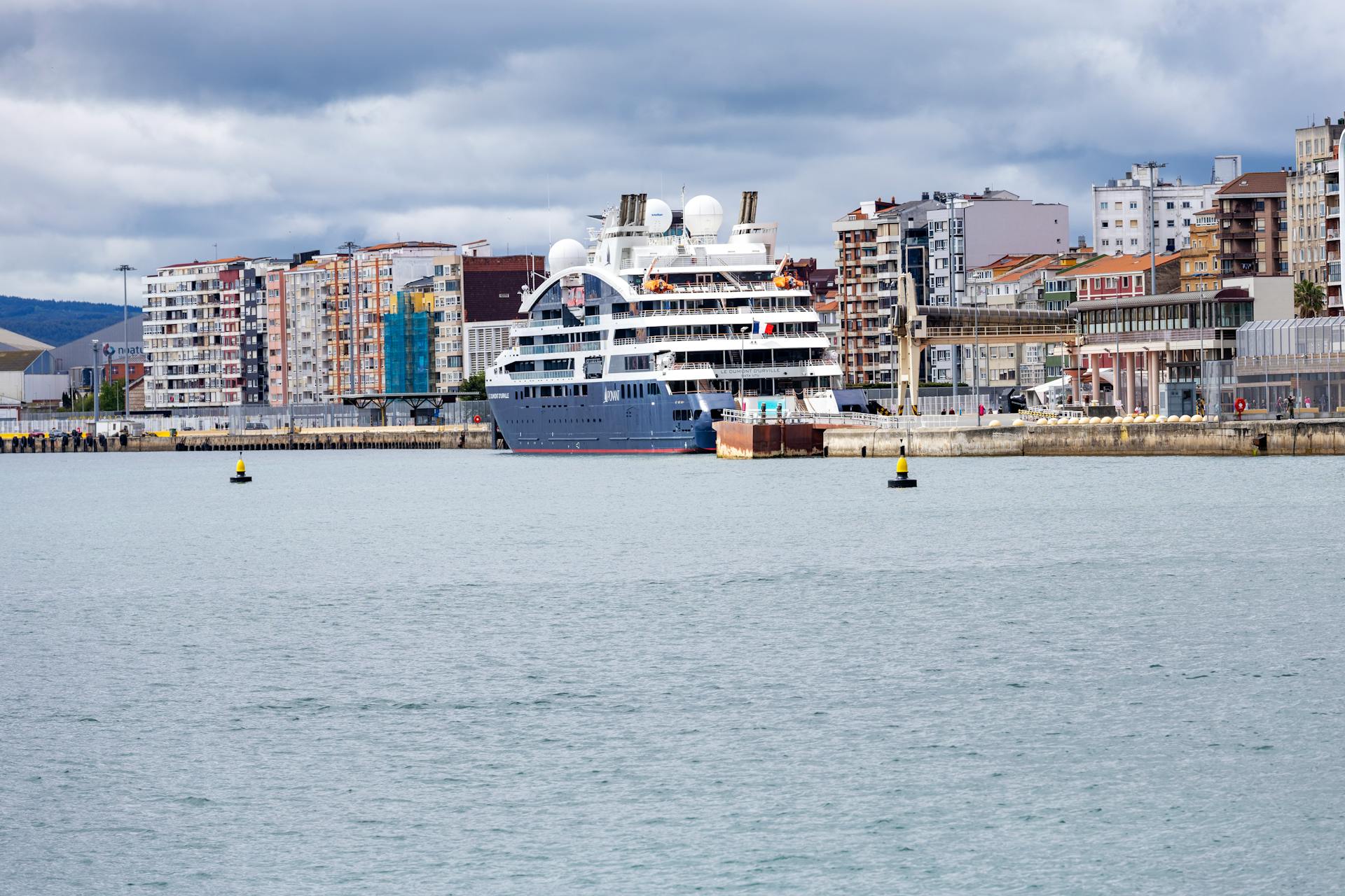 Large cruise ship docked at a city port with a scenic urban backdrop on a cloudy day.