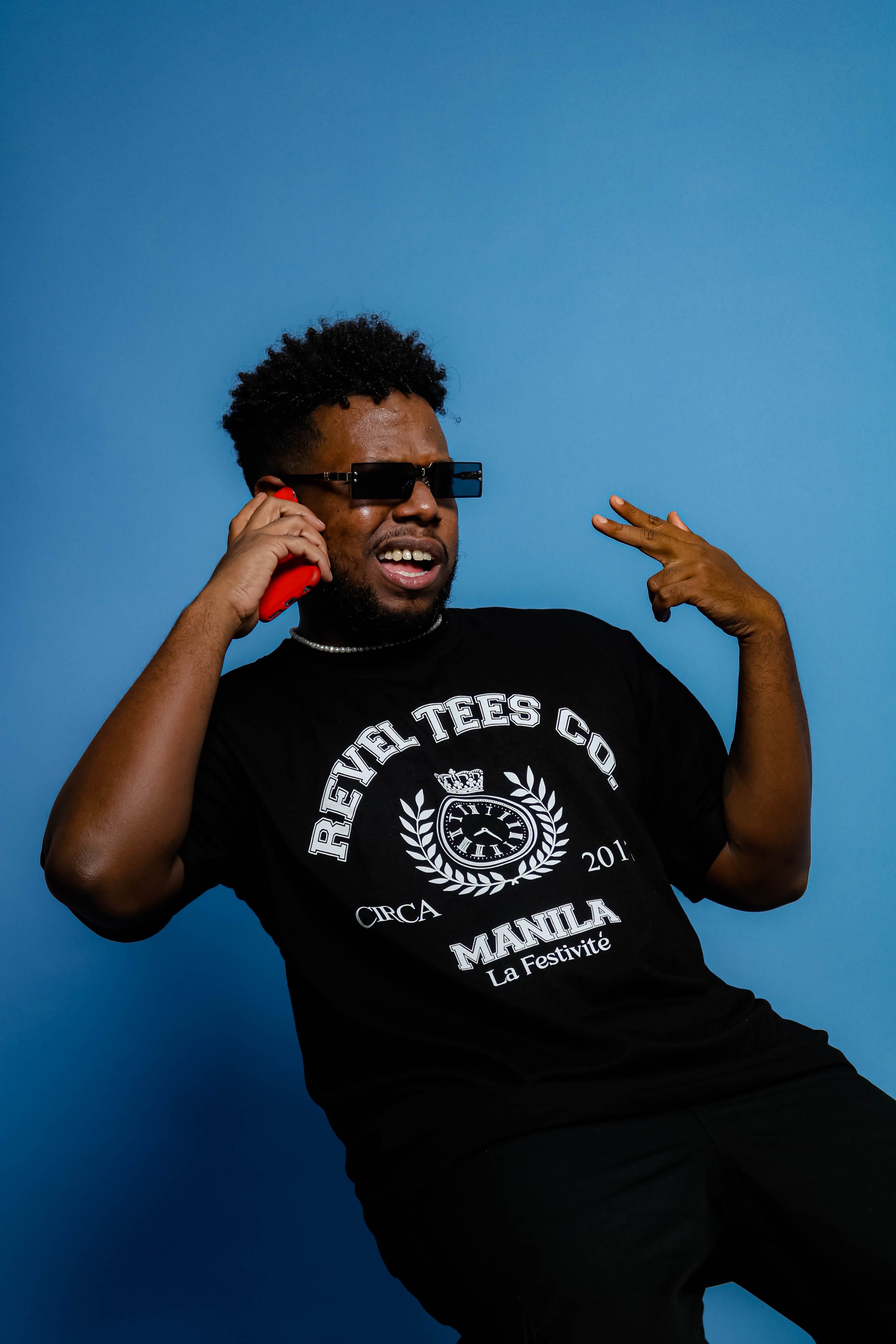 A stylish man in sunglasses chats on a red smartphone against a blue backdrop in a studio.