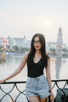 A stylish woman in denim shorts poses by the waterfront with an urban cityscape in the background.