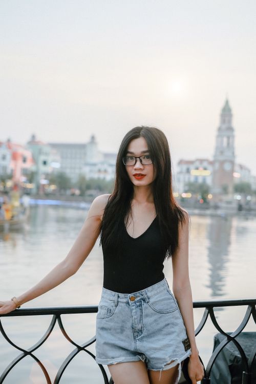 Free A stylish woman in denim shorts poses by the waterfront with an urban cityscape in the background. Stock Photo