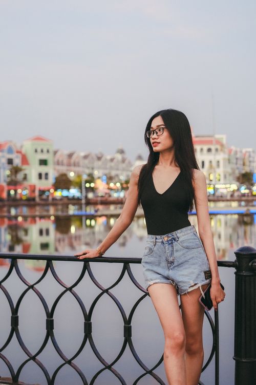 Free Trendy young woman poses by a scenic waterfront promenade at dusk, creating a relaxed vacation vibe. Stock Photo