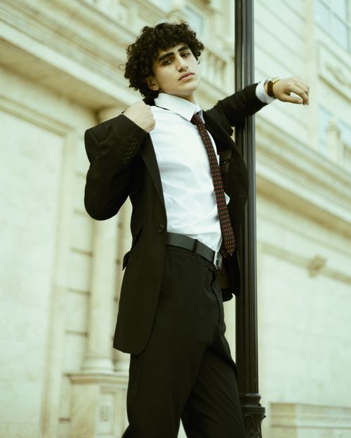 Free Young man in a black suit with a necktie, leaning against a lamppost outdoors, exuding elegance. Stock Photo