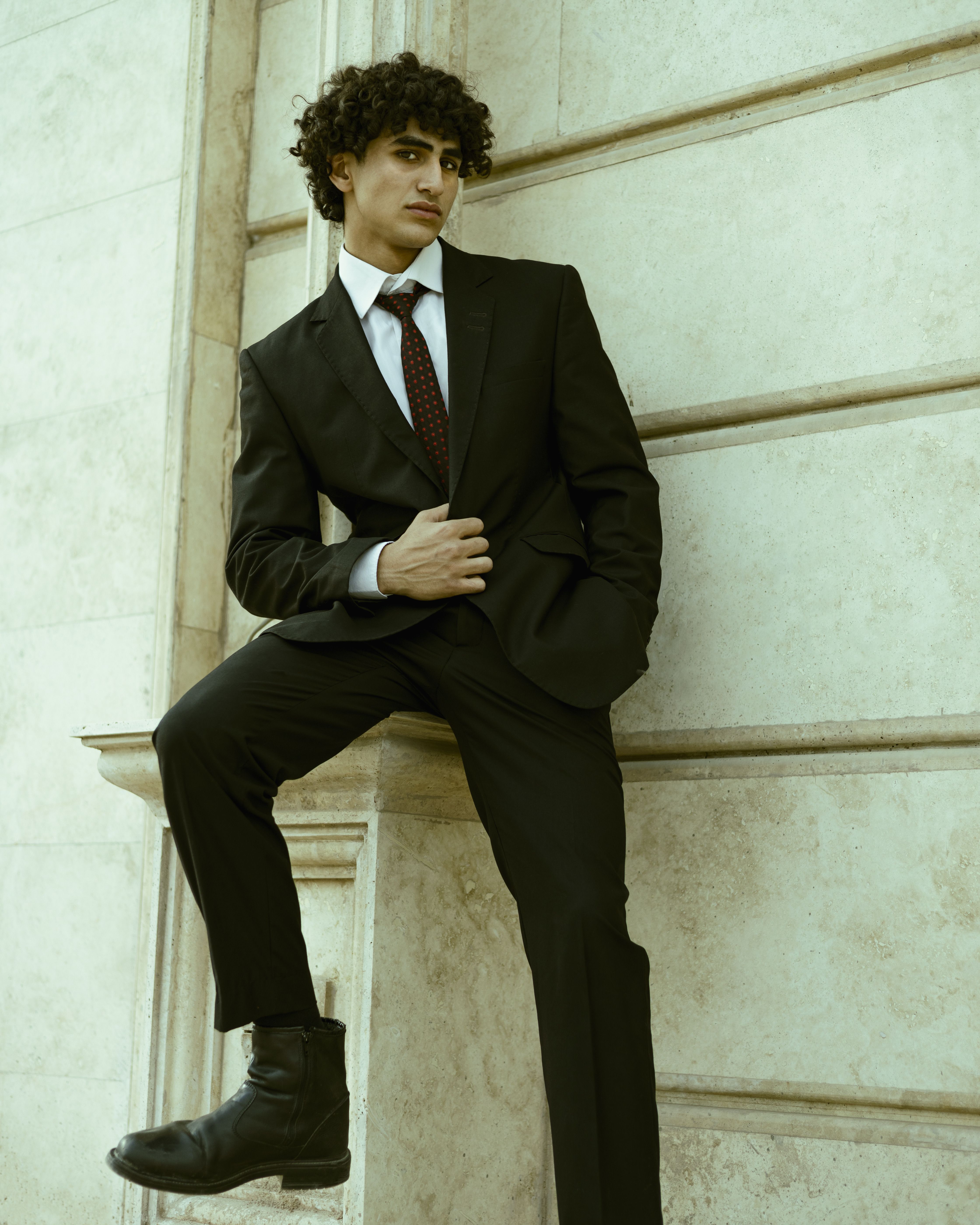 Fashionable young adult male in a black suit leans casually against a white stone wall.