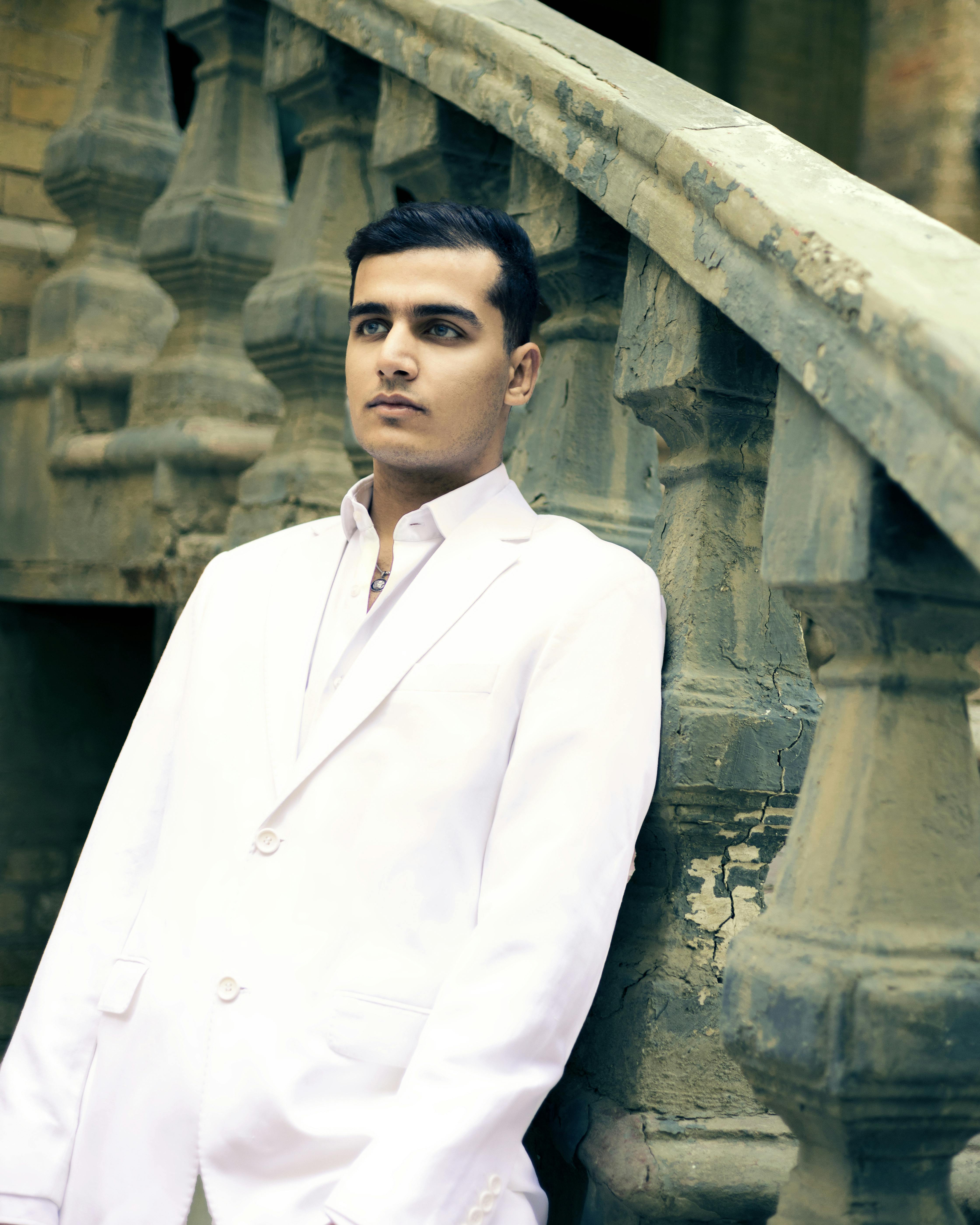 A stylish young man in a white suit poses elegantly against ornate urban stairs, showcasing fashion trends.
