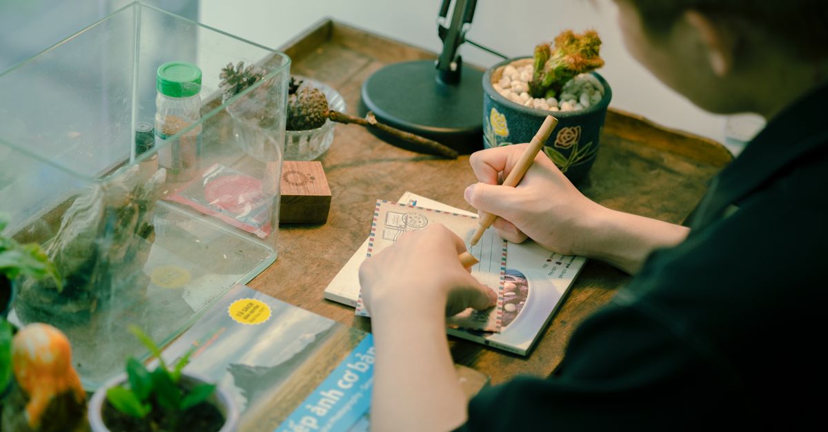 Close-up of a person writing a postcard on a wooden desk with plants and decor.
