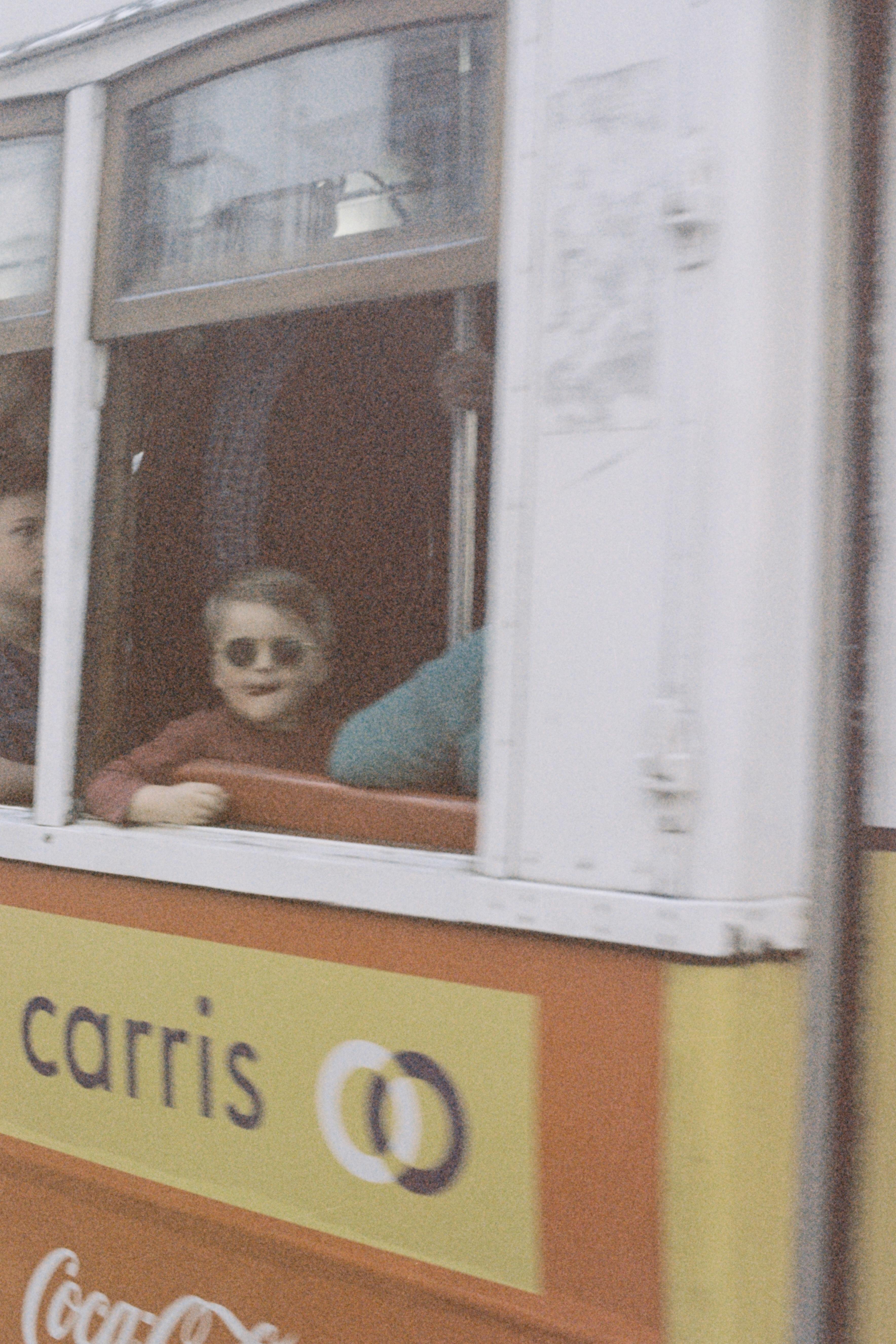 A young boy wearing sunglasses enjoys a tram ride in Lisbon, captured through the window for a candid moment.